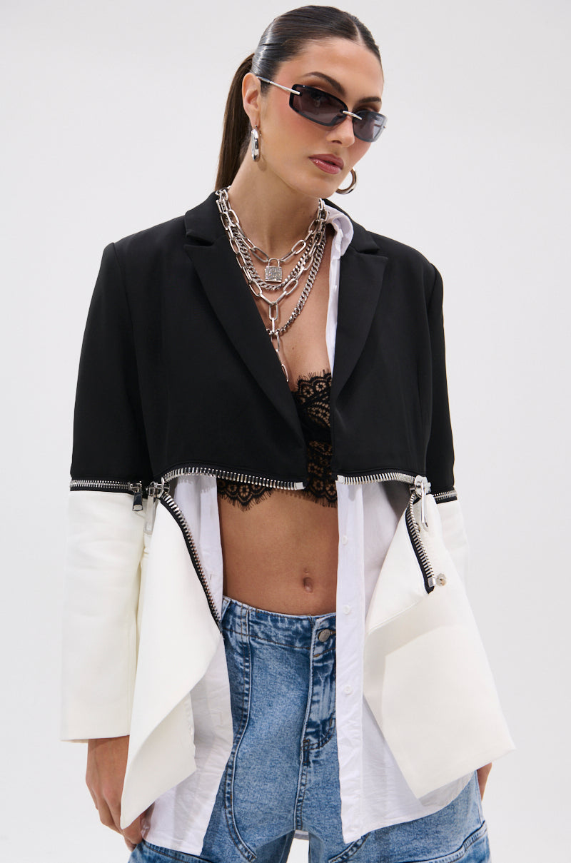A woman wearing sunglasses, layered necklaces, the DON'T TRIP ZIPPER BLAZER over a lace bralette and high-waisted blue jeans stands against a plain white background.