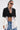 A woman wearing sunglasses, layered necklaces, the DON'T TRIP ZIPPER BLAZER over a lace bralette and high-waisted blue jeans stands against a plain white background.