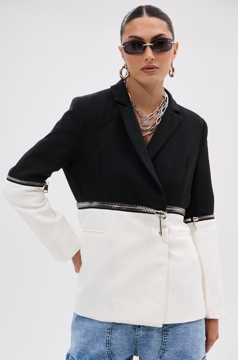 Wearing the DON'T TRIP ZIPPER BLAZER with black sunglasses, silver hoop earrings, layered necklaces, and light blue jeans, a person poses with one hand on their hip against a plain background.