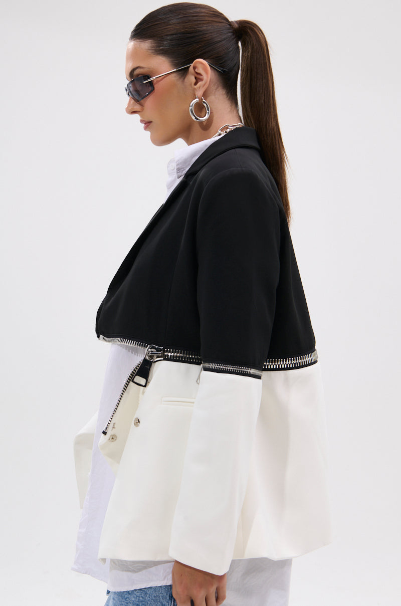 A woman with a ponytail wears sunglasses, large hoop earrings, and the DON'T TRIP ZIPPER BLAZER—a striking piece featuring a black top half and white bottom half. She stands sideways against a plain background.