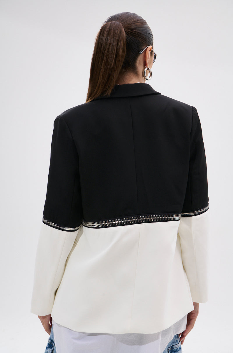 A person with long brown hair in a ponytail wears sunglasses and the DON'T TRIP ZIPPER BLAZER, a dual-tone black and white piece with decorative trim dividing the colors, seen from the back.