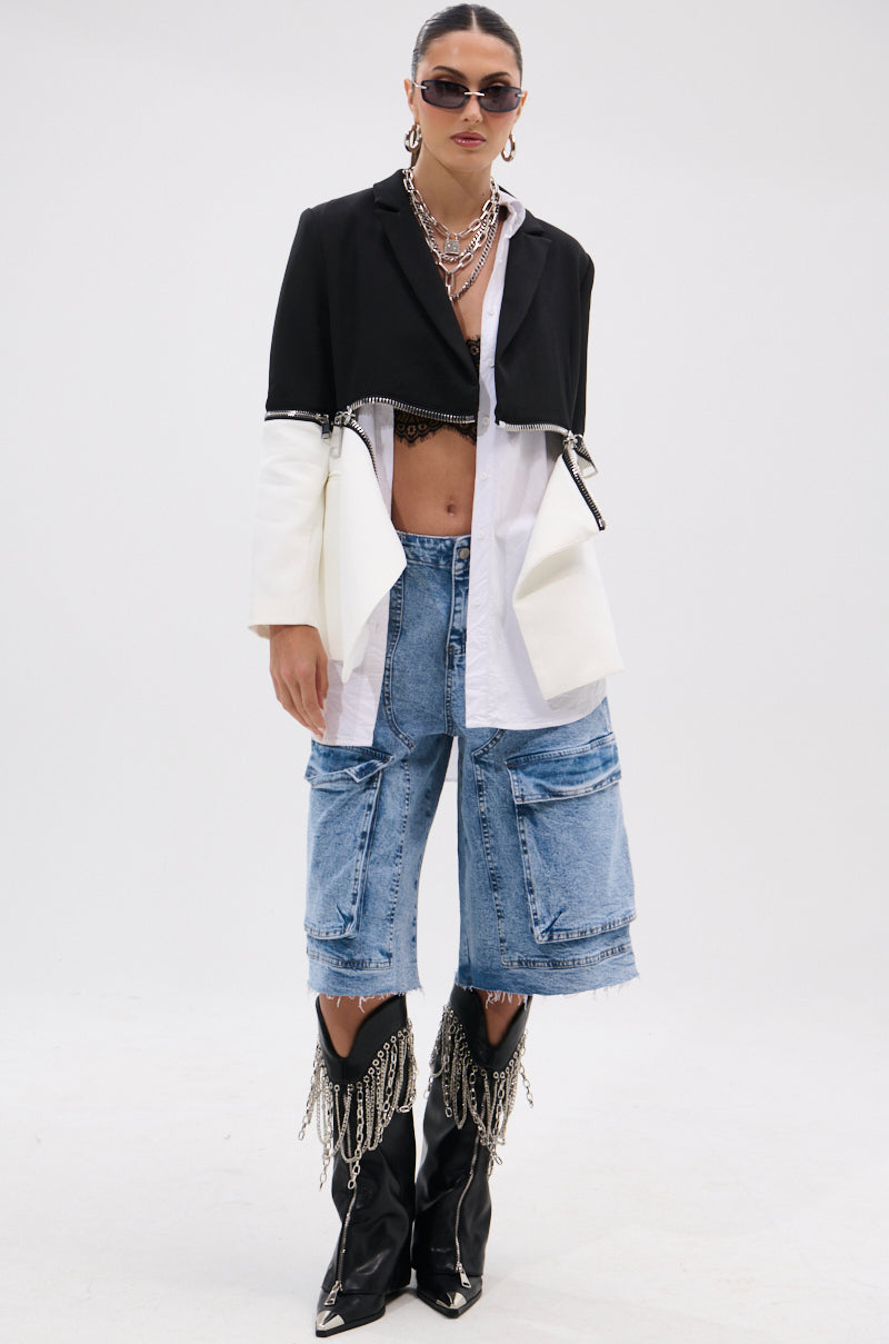 A person models the DON'T TRIP ZIPPER BLAZER—a cropped, dual-tone black-and-white piece—over a white shirt, paired with long jean shorts and accessories like black chain-detailed boots, layered necklaces, and sunglasses against a plain white background.