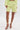 A person wears the SMELL THE FLOWERS BERMUDA SHORT in light yellow with a matching blazer and gold strappy heels, standing on a white background. A box-shaped handbag peeks out behind them, adding to the chic pastel outfit.