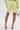 Wearing the SMELL THE FLOWERS BERMUDA SHORT and a matching blazer, paired with white ankle-tie heeled sandals and a small transparent handbag, someone stands facing away for a chic pastel look.