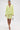 A woman in the SMELL THE FLOWERS BERMUDA SHORT, a matching yellow-green blazer, strappy heels, and a box-shaped handbag stands against a white background for a chic pastel look.