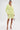 A woman stands confidently in the SMELL THE FLOWERS BERMUDA SHORT paired with a matching light yellow blazer, gold strappy heels, and a metallic mini handbag. Her dark hair is pulled back against a plain white background.