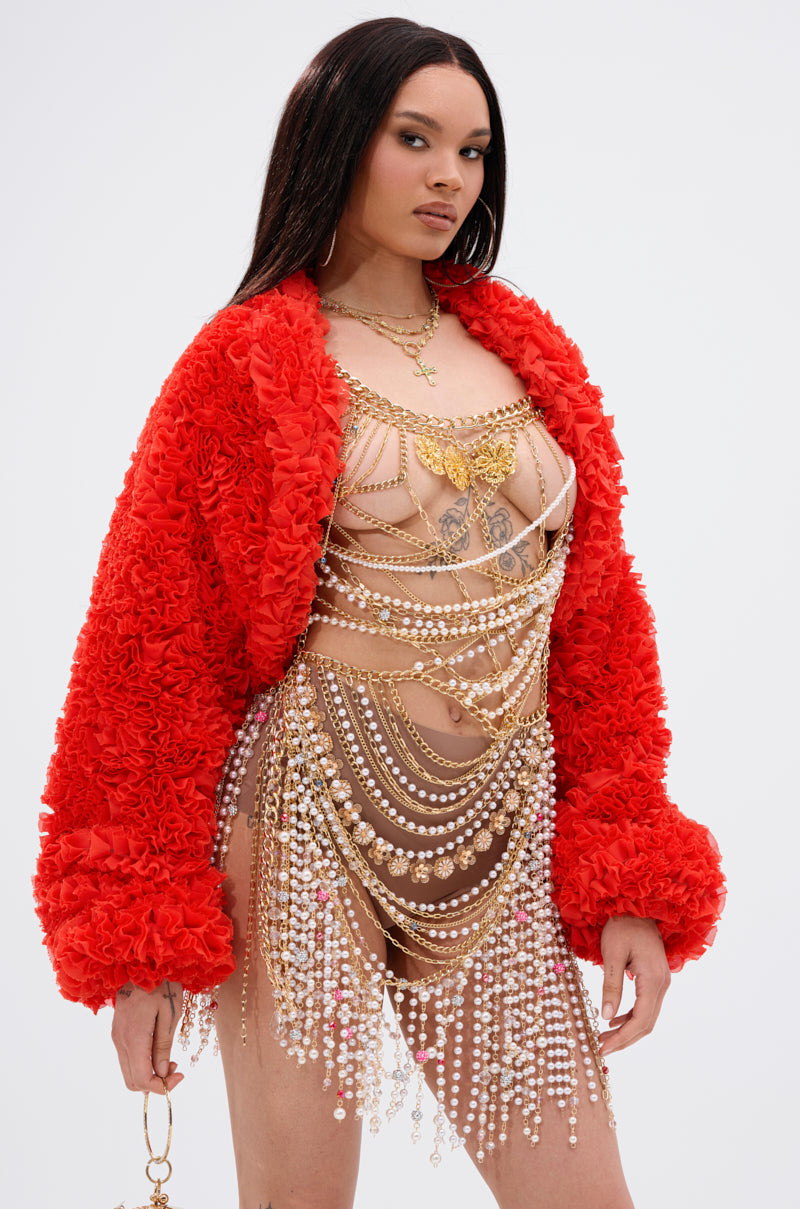 A woman wears the FIRED UP CHIFFON SHRUG—a bright red textured jacket with exaggerated sleeves—paired with an ornate gold and pearl chain dress, standing against a plain white background.