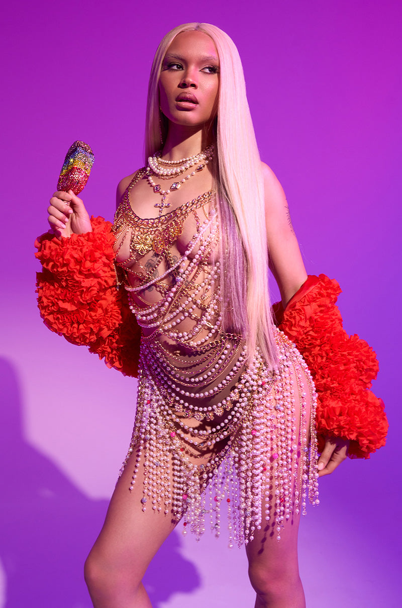 Wearing the FIRED UP CHIFFON SHRUG, a person with long blonde hair poses against a purple background, styled with layered pearls, jewels, metallic accessories, and holding a colorful bejeweled popsicle.