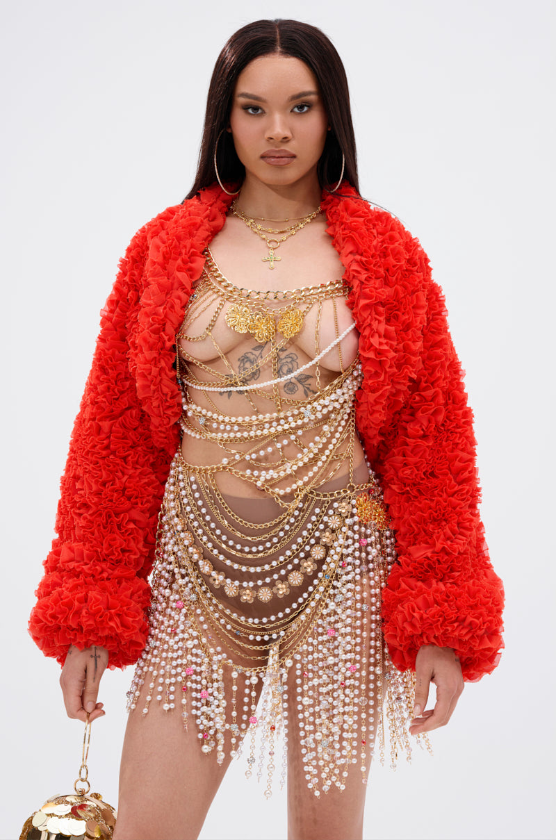 Wearing the FIRED UP CHIFFON SHRUG—a textured red jacket with exaggerated sleeves—a woman layers gold chains and pearl body jewelry over a nude outfit, accessorized with a gold purse, hoop earrings, and straight hair against a plain background.