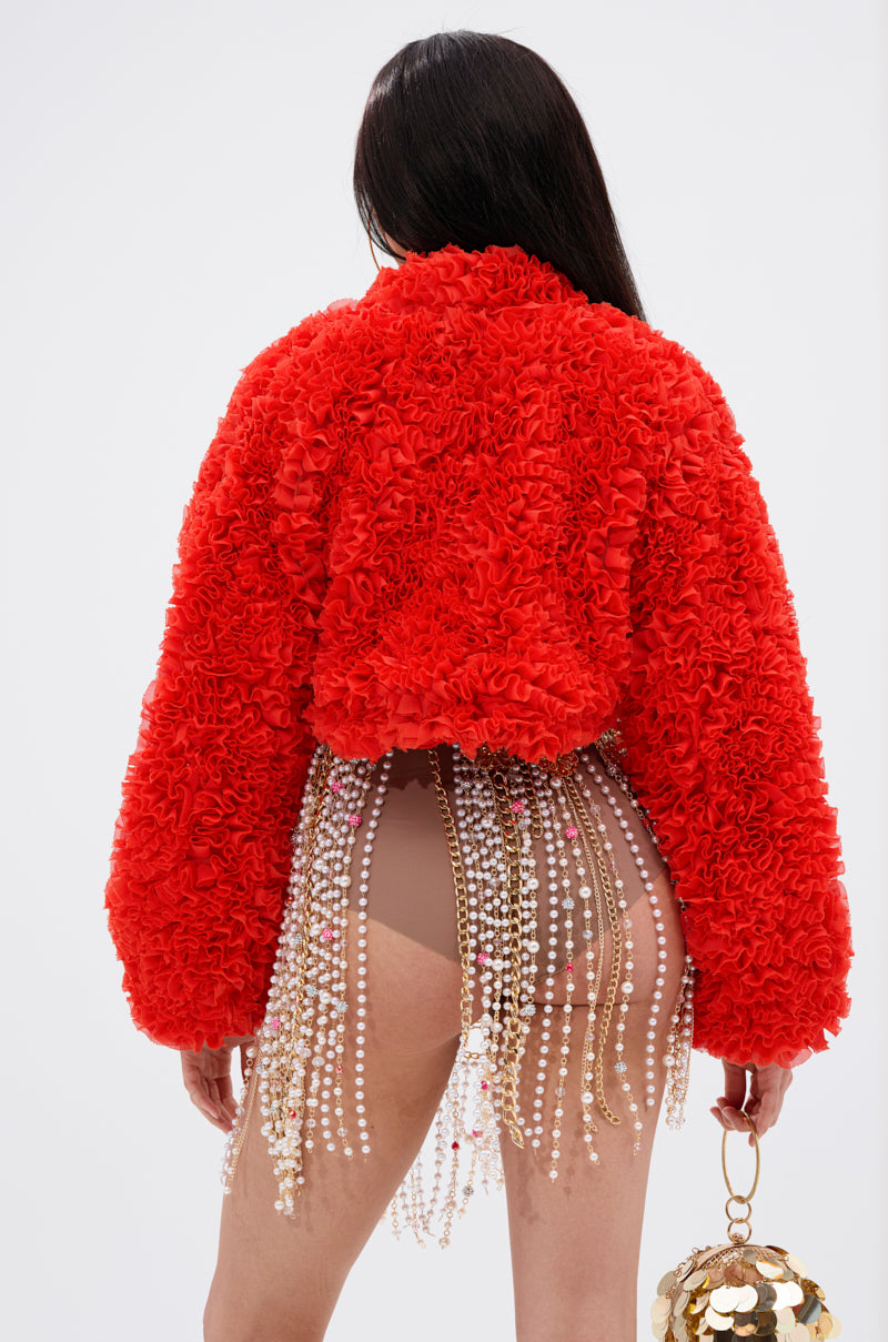 A person faces away, wearing the FIRED UP CHIFFON SHRUG—a bright red, textured, ruffled jacket with exaggerated sleeves—paired with a sheer beaded skirt and a gold reflective spherical handbag against a plain white background.