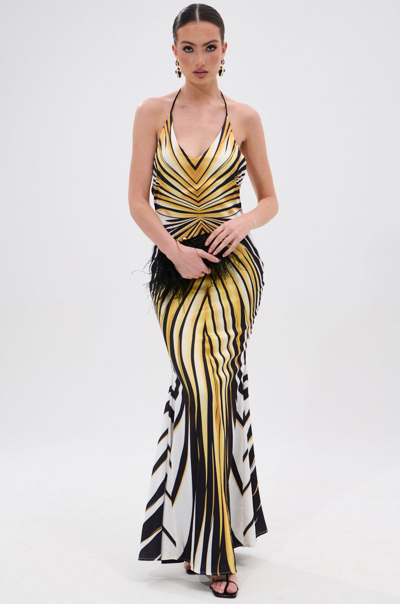 A woman stands against a white background wearing the COVER STAR PRINTED SATIN MIDI DRESS, a long, sleeveless halter-neck dress with bold yellow, white, and black geometric stripes, styled with a black clutch and black heels.