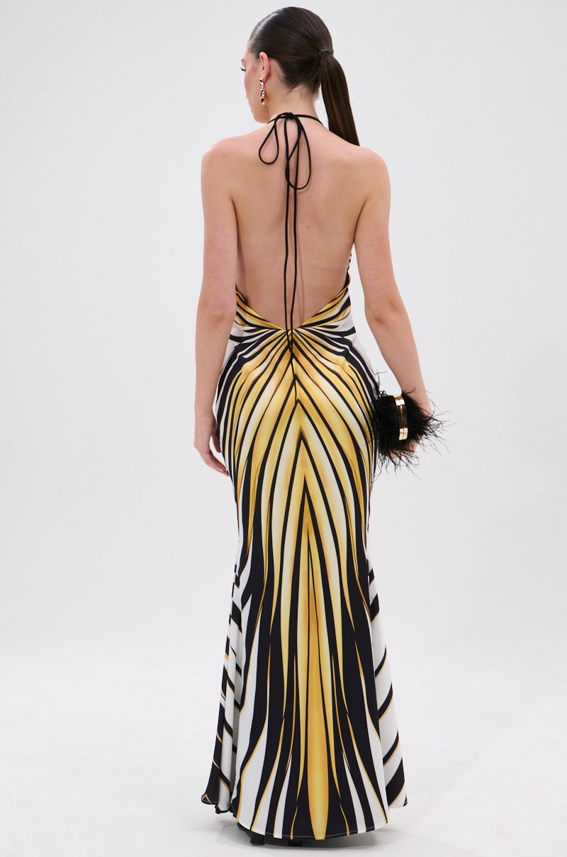 A woman faces away in the COVER STAR PRINTED SATIN MIDI DRESS, a backless halter-neck gown with a yellow, black, and white geometric print and mermaid silhouette. She holds a black feathered clutch and wears her hair in a sleek ponytail.