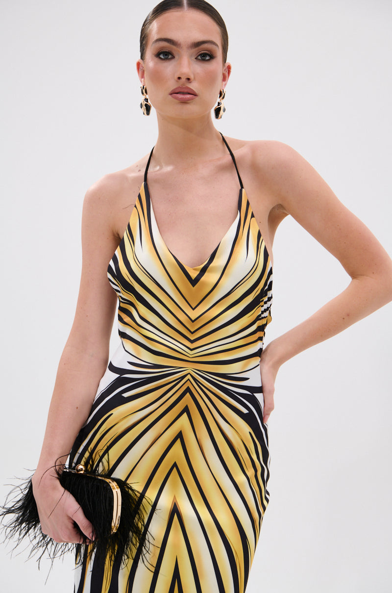 A woman wears the COVER STAR PRINTED SATIN MIDI DRESS, a sleeveless gold and black geometric halter with a mermaid silhouette, posing against a plain background with a feather-accented black clutch and gold earrings.