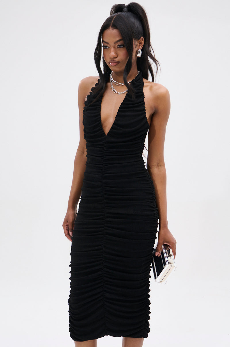 A woman poses against a plain background in the BODY ODDY MIDI DRESS IN BLACK, accessorized with a small black and white clutch. Wearing earrings, a necklace, and her hair styled in a high ponytail, she completes the chic ensemble.