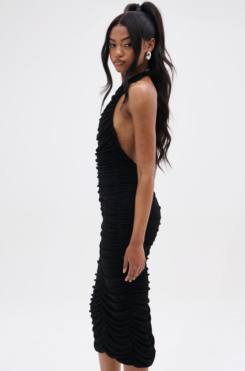 A woman with long black hair stands sideways wearing the BODY ODDY MIDI DRESS IN BLACK, a sleeveless, ruched mesh dress that hugs her figure like a classic little black dress. Her high ponytail and earrings add elegance as she gazes at the camera against a plain white background.