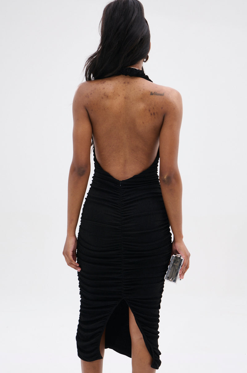 A person with long hair is wearing the BODY ODDY MIDI DRESS IN BLACK, featuring an alluring backless design. They stand against a plain background, elegantly holding a small rectangular clutch in their left hand.