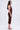 A woman in profile wears the BODY ODDY MIDI DRESS IN BROWN, a sleeveless and ruched midi dress, paired with high heels. She has long, wavy hair and poses elegantly against a plain, light background.