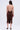 A person, with wavy hair cascading freely, stands facing away from the camera to showcase the BODY ODDY MIDI DRESS IN BROWN. This ruched midi dress features a halter neckline and a bottom slit, paired with high-heeled sandals against a simple, light-colored backdrop.