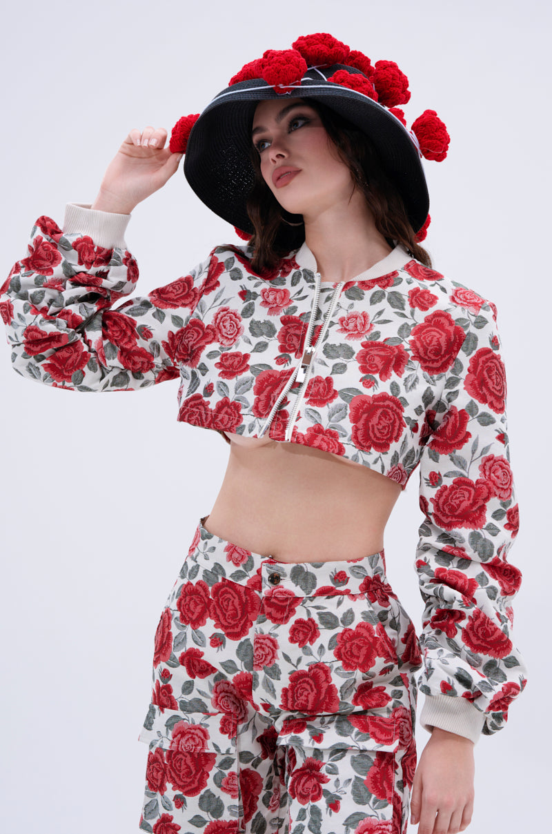 A woman models the FULL OF ROSES CROPPED BOMBER, a rose-print floral jacket paired with a matching wide-brimmed hat with red pom-poms, set against a plain light background.