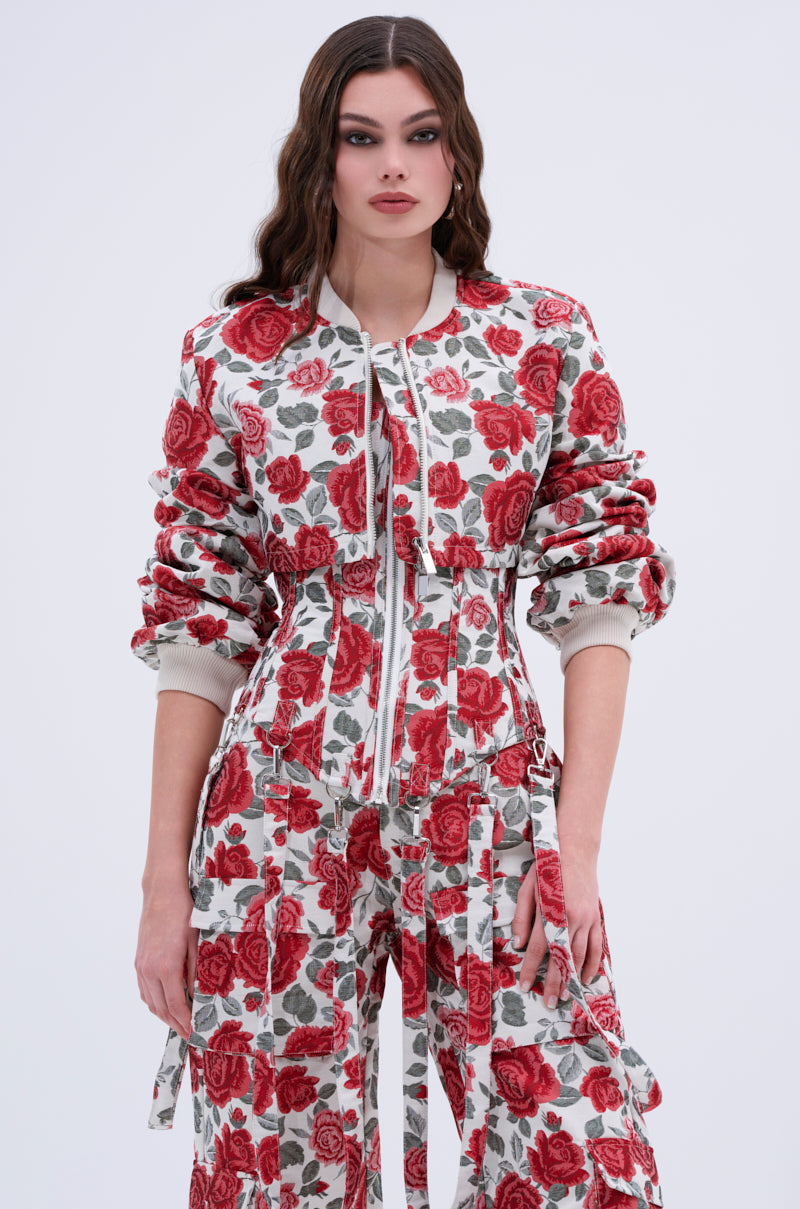 A woman with long wavy brown hair wears the FULL OF ROSES CROPPED BOMBER, a rose print floral jacket featuring bold red roses and green leaves on white, paired with wide-leg pants, embracing streetwear against a plain light background.