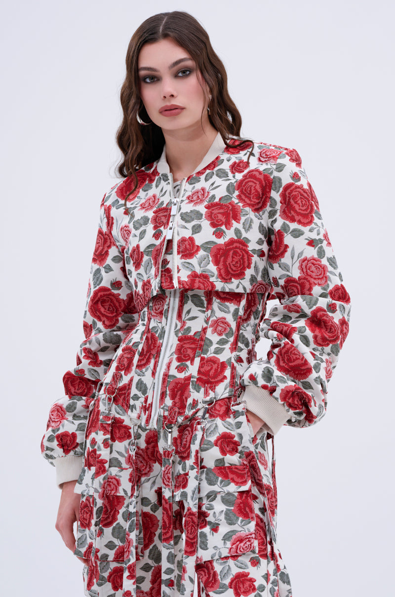A woman with long wavy brown hair and hoop earrings models the FULL OF ROSES CROPPED BOMBER and matching high-waisted pants with zipper details, embodying chic streetwear style against a plain background.