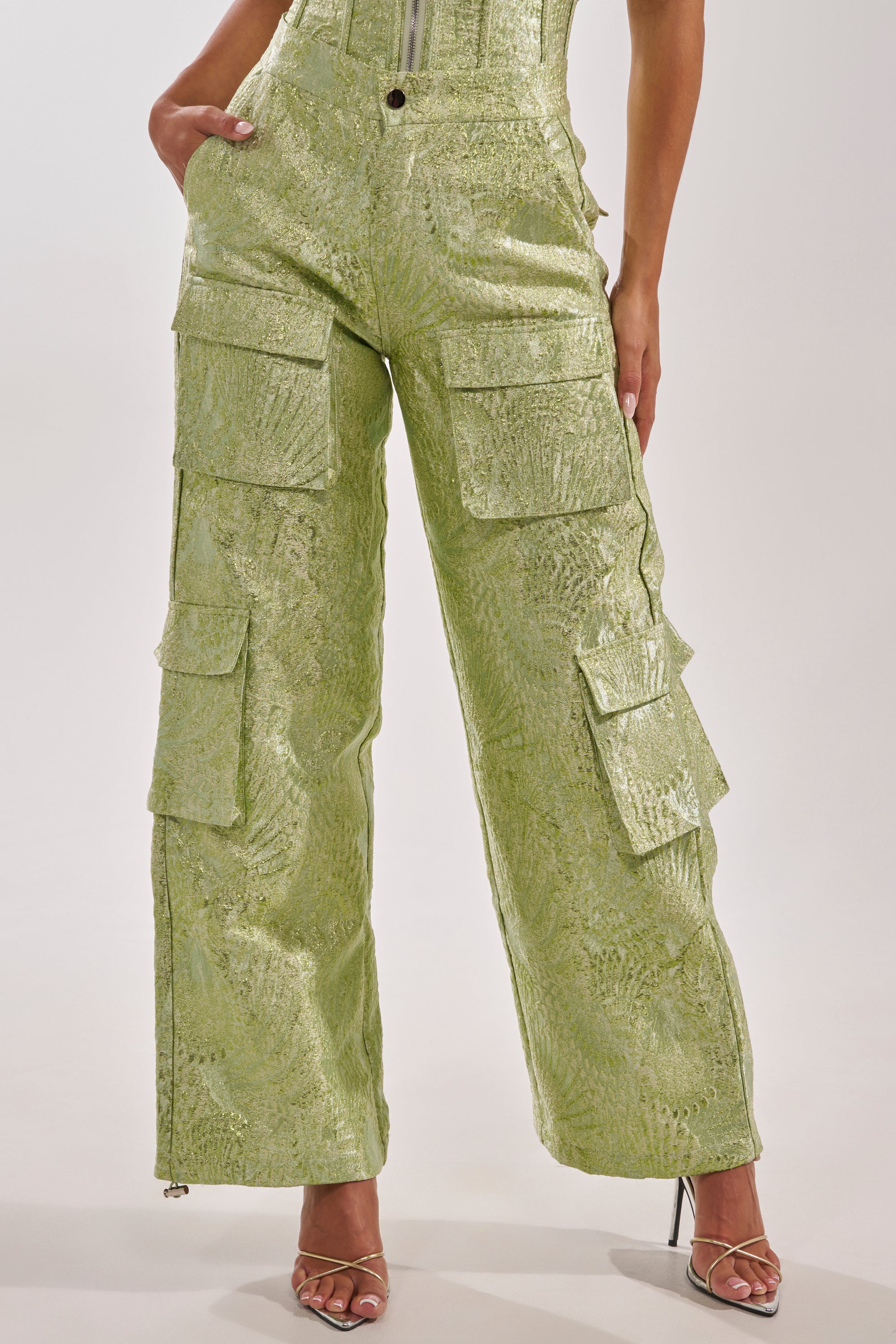 Wearing ENVY ME BROCADE PANT—high-waisted, light green shimmer cargo pants with large pockets—a person stands confidently in strappy heels, one hand in a pocket. The image is waist-cropped, so the face isn't visible. 2