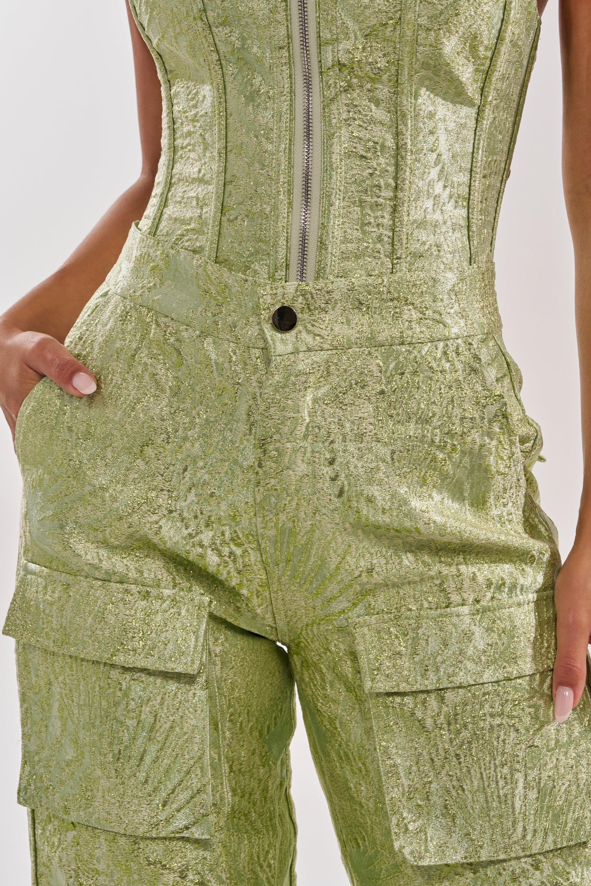 Close-up of a person wearing the ENVY ME BROCADE PANT in shiny, textured green. Their left hand is in their pocket, with only the torso and upper legs visible. 5