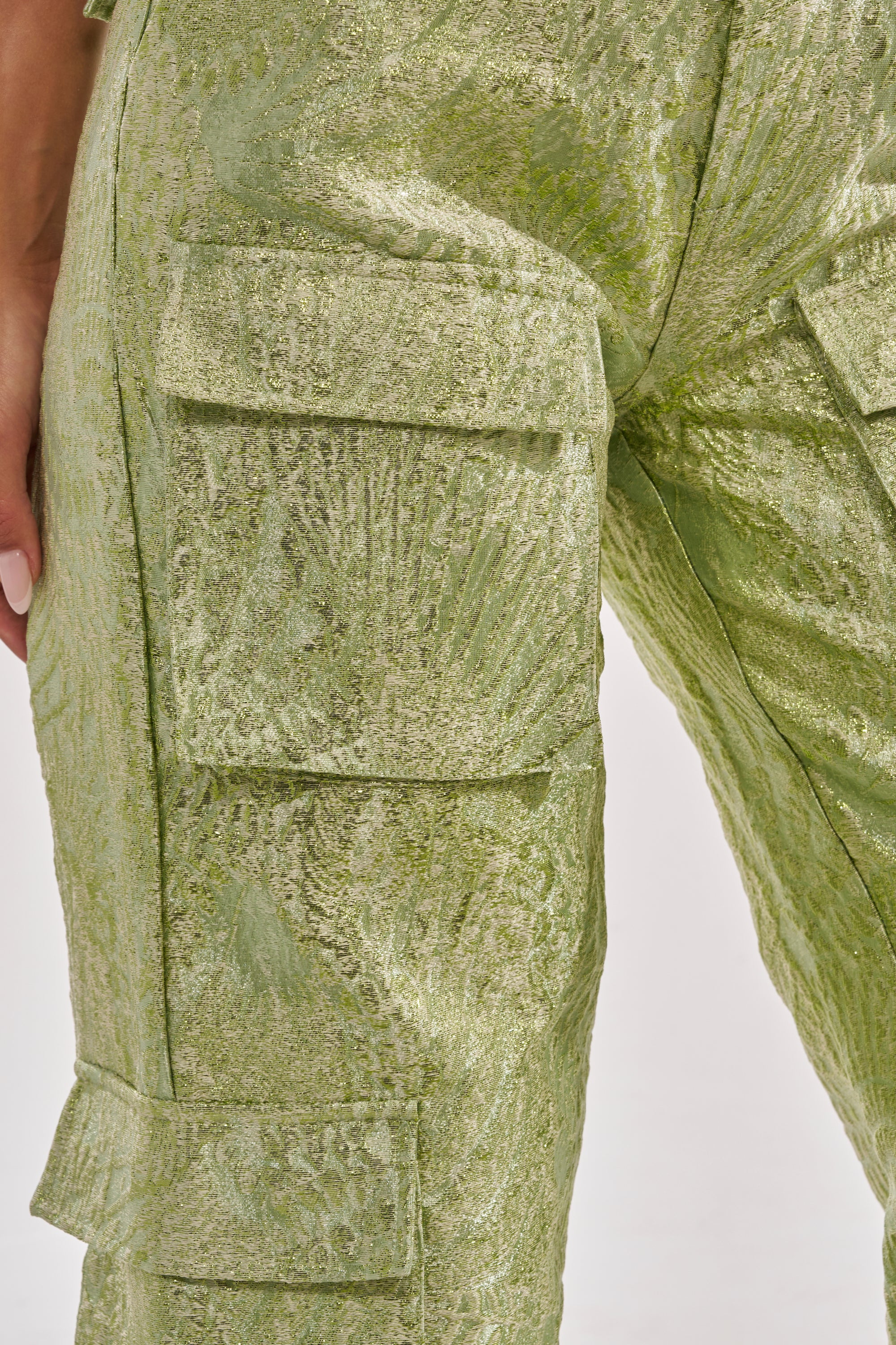 Close-up of a person wearing the ENVY ME BROCADE PANT, featuring shimmering green patterned fabric and flap pockets—a striking statement piece for streetwear enthusiasts. Manicured nails are visible at the edge. 11