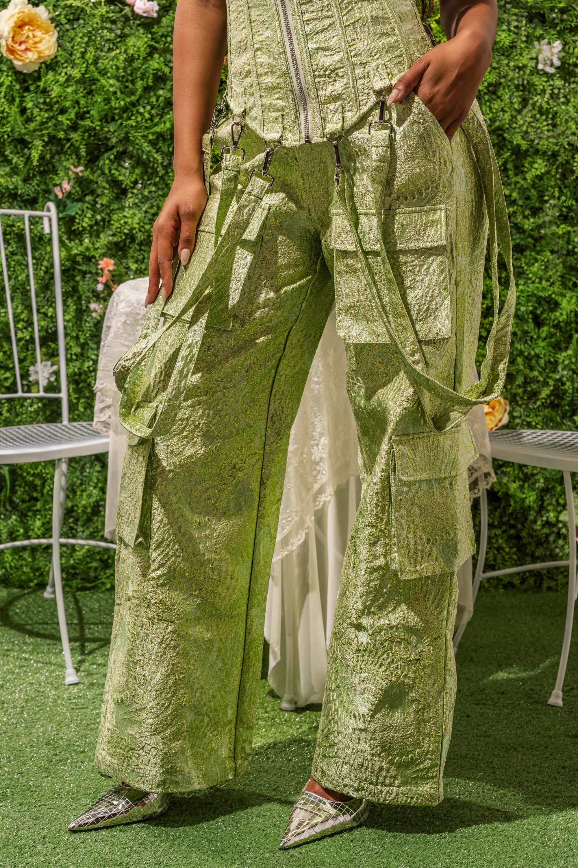 A person stands outdoors in the ENVY ME BROCADE PANT, styled as streetwear with a matching textured corset top. The shimmering green outfit pops against white chairs and a lush green floral backdrop. 0