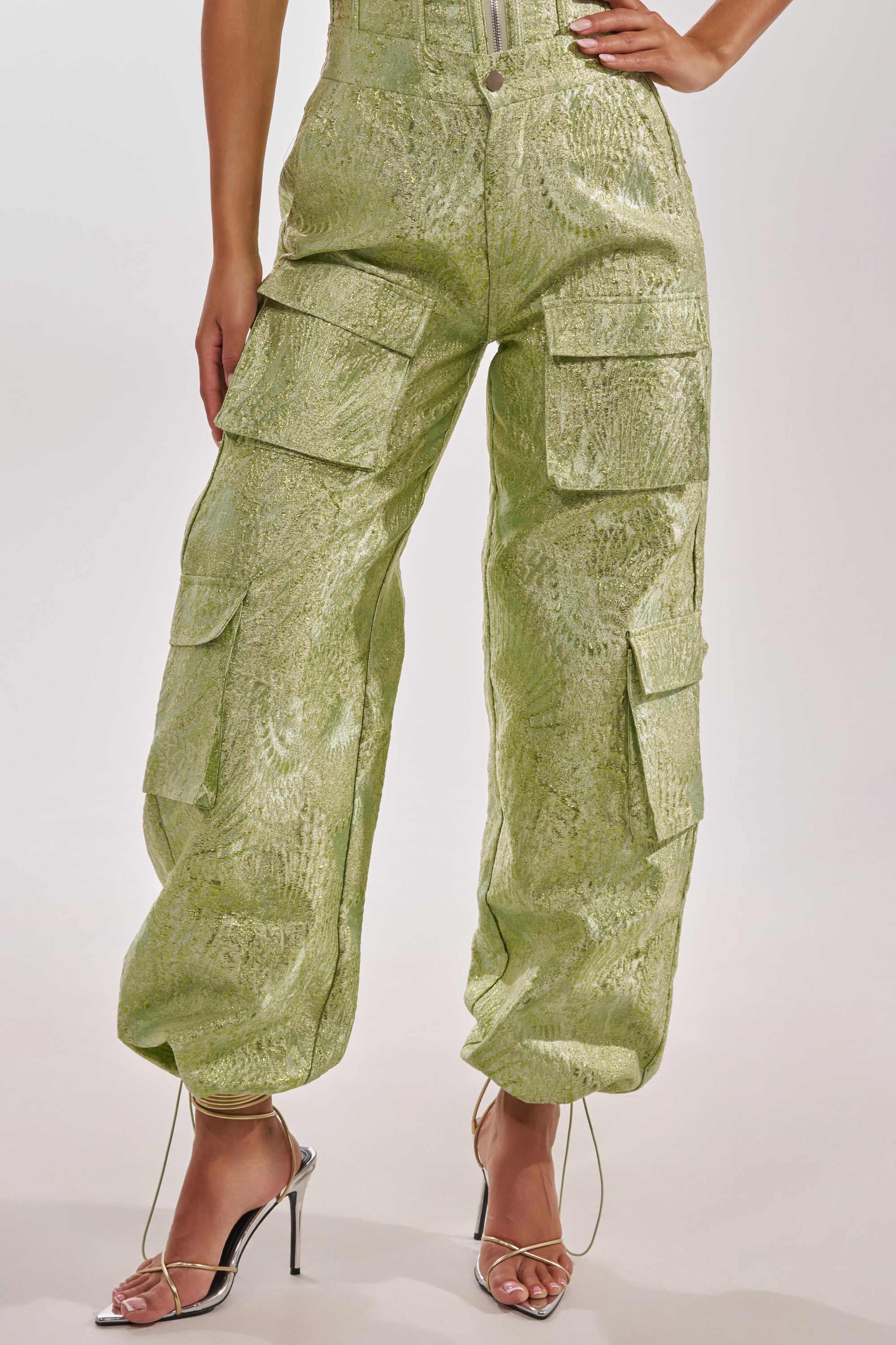 A person models the ENVY ME BROCADE PANT, featuring multiple pockets and a zipper front, styled with silver strappy high-heeled sandals against a plain background. 3