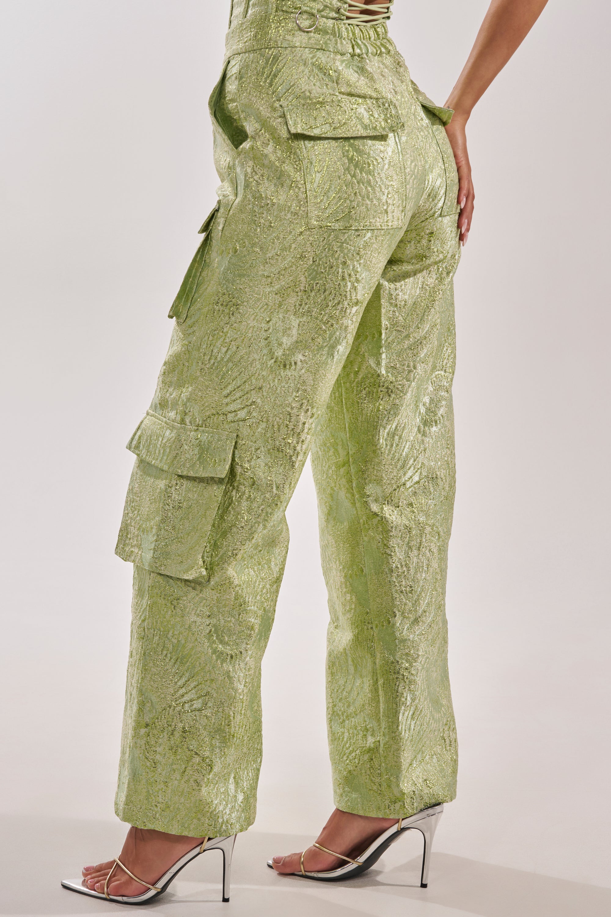 A person models the ENVY ME BROCADE PANT—high-waisted, shiny light green cargo pants with side pockets—paired with silver heels, shown from the side and back against a plain background. 4