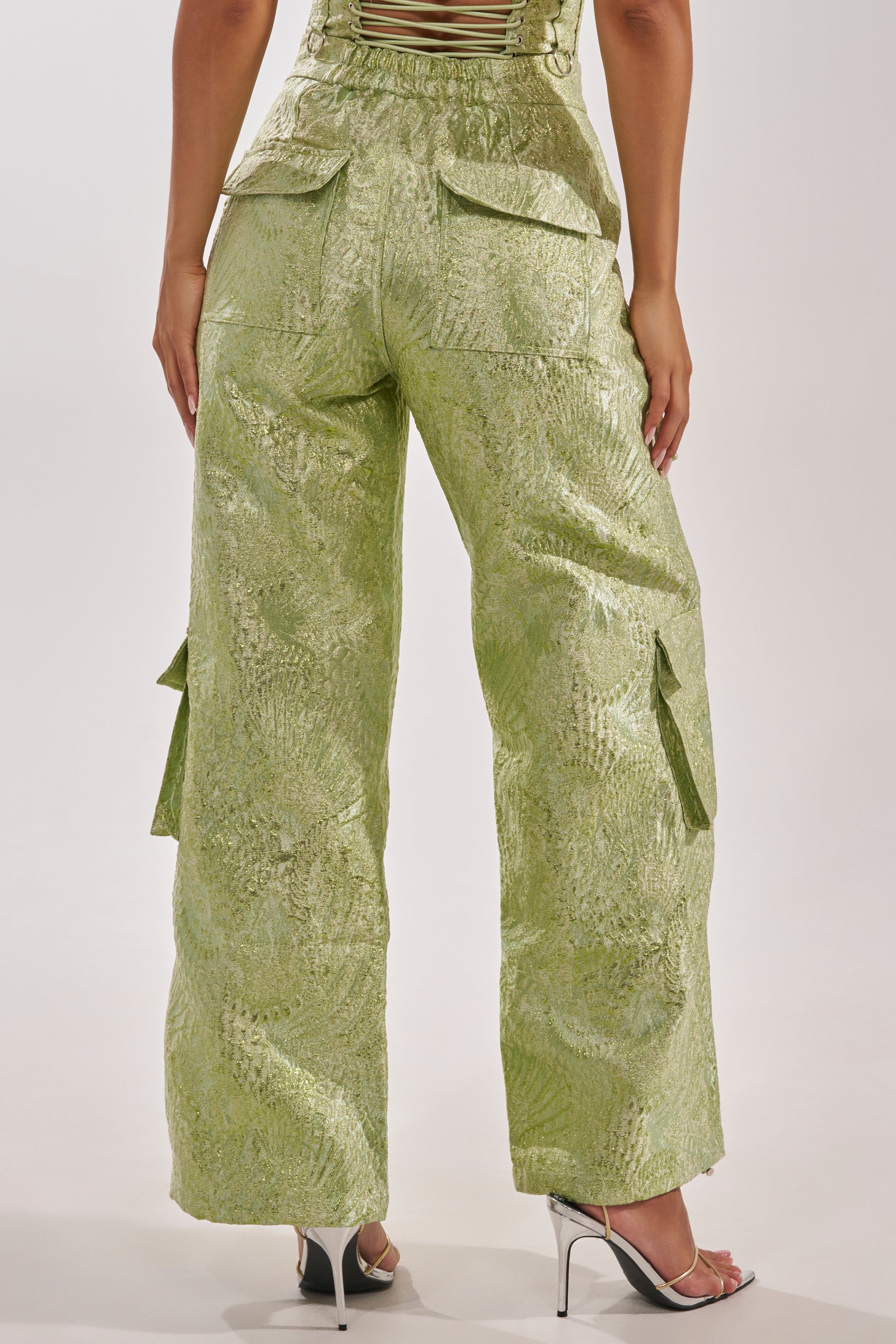 A person shown from the back wears ENVY ME BROCADE PANT—shiny, light green shimmer cargo pants with a textured finish and flap pockets—paired with high heels. 6