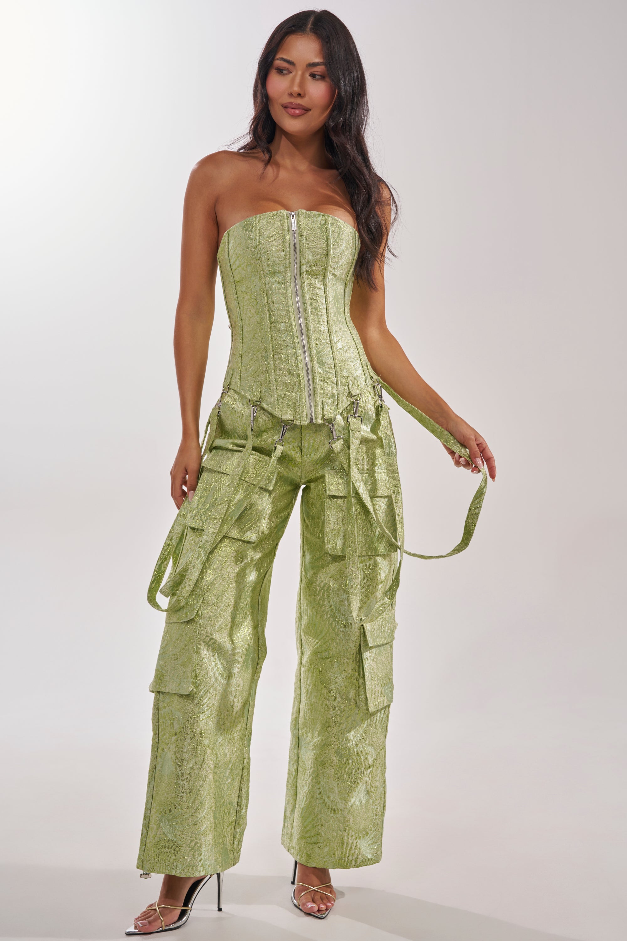 Woman wears the ENVY ME BROCADE PANT—light green cargo pants with brocade texture, multiple pockets, and hanging straps—paired with a matching strapless corset top and clear heels, standing against a light background. 10