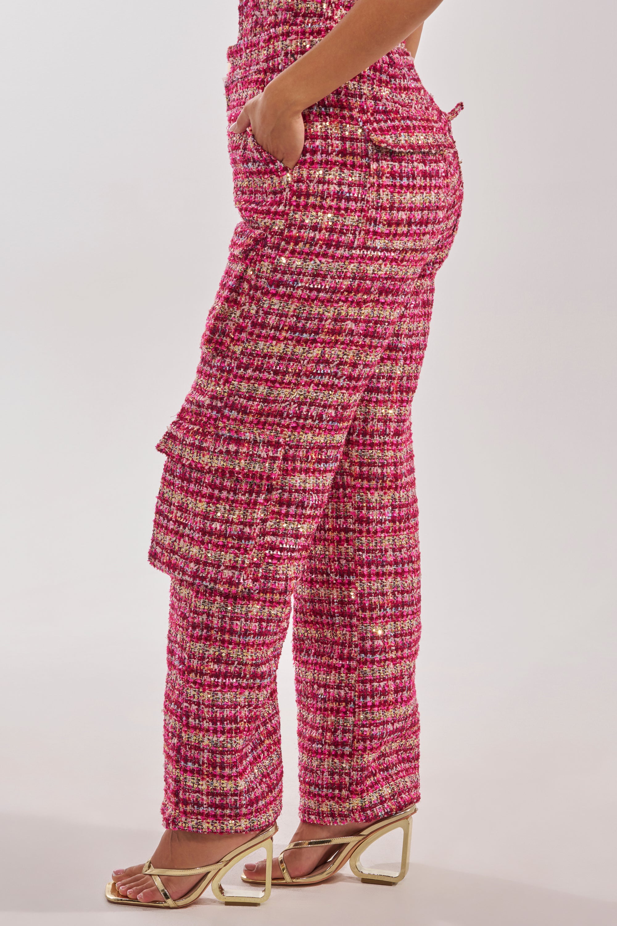A person is wearing the ITS TIME TWEED TROUSER in pink and white, featuring a high-rise waist, side pockets, and layered flap details. Styled with metallic gold open-toe heels, the photo highlights the straight-leg silhouette from a side angle. 2