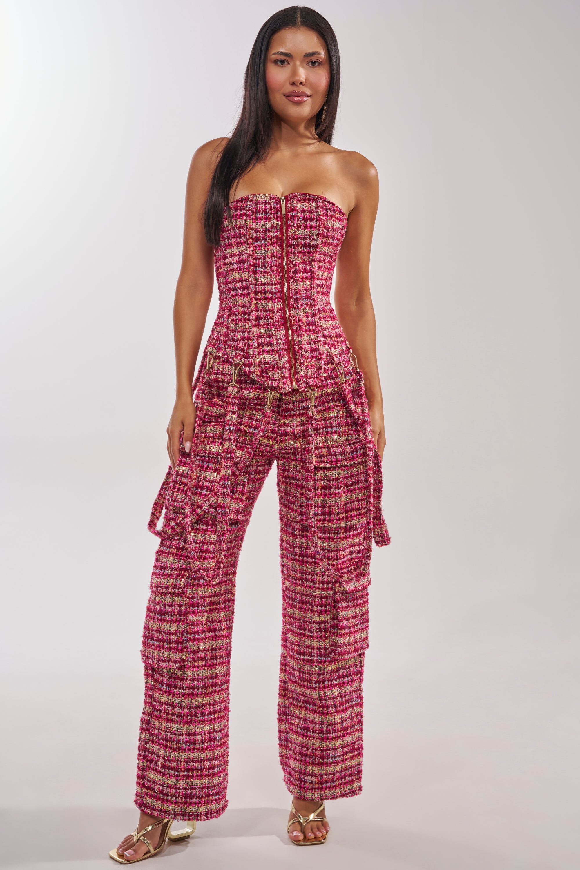 A woman wears the ITS TIME TWEED TROUSER, a high-rise, pink and red fitted tweed pant with metallic threads and flared legs, paired with gold open-toe heels against a plain light background. 4