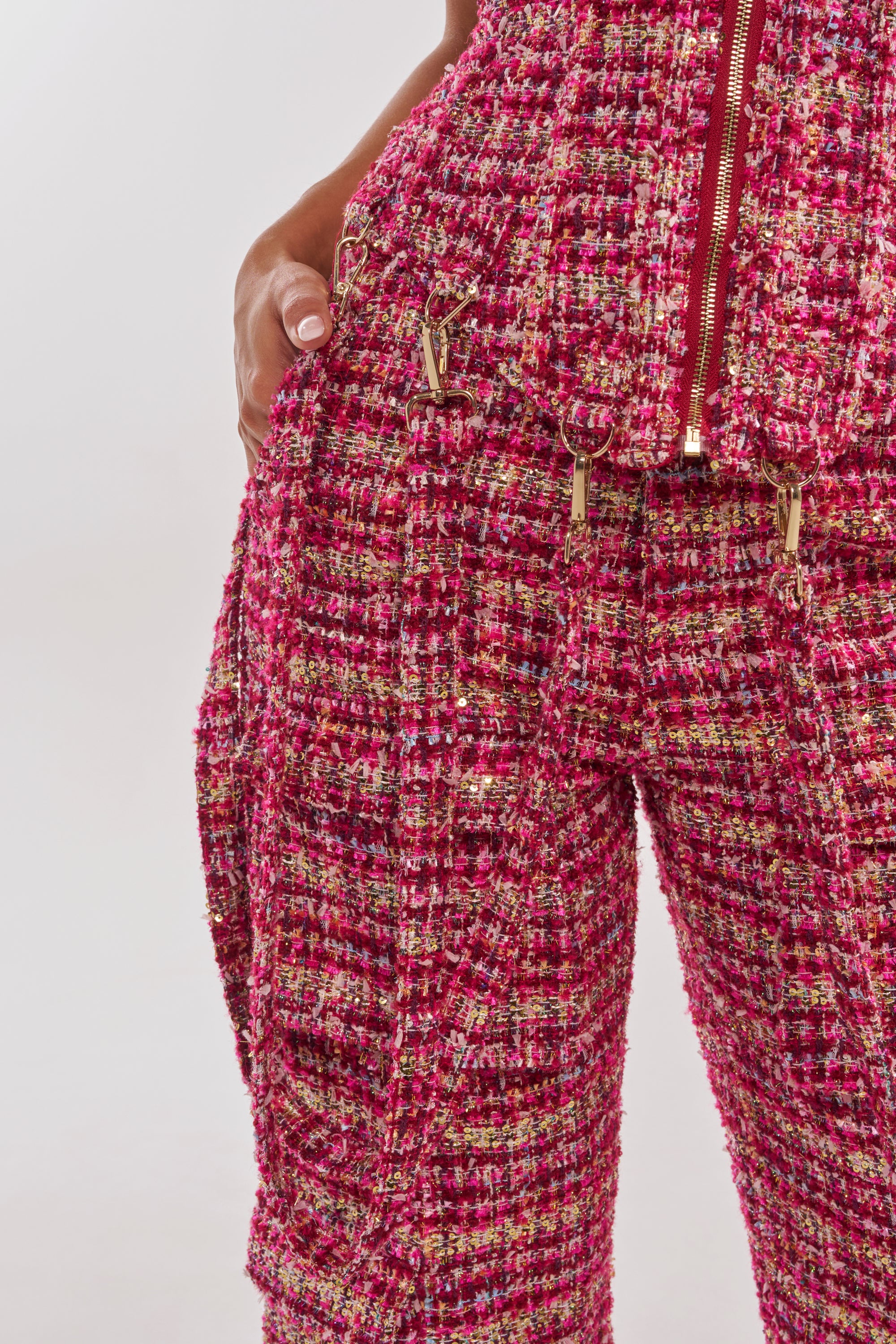 A close-up of a person wearing the ITS TIME TWEED TROUSER—textured pink, gold, and white plaid tweed with metallic threads—paired with a matching jacket featuring a red zipper, gold hardware, and one hand in the pocket. 6