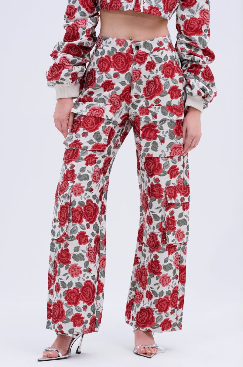 A person wears the FULL OF ROSES TROUSER—high-waisted with a bold red rose print—paired with a matching cropped jacket and silver open-toe heels, standing against a plain white background.