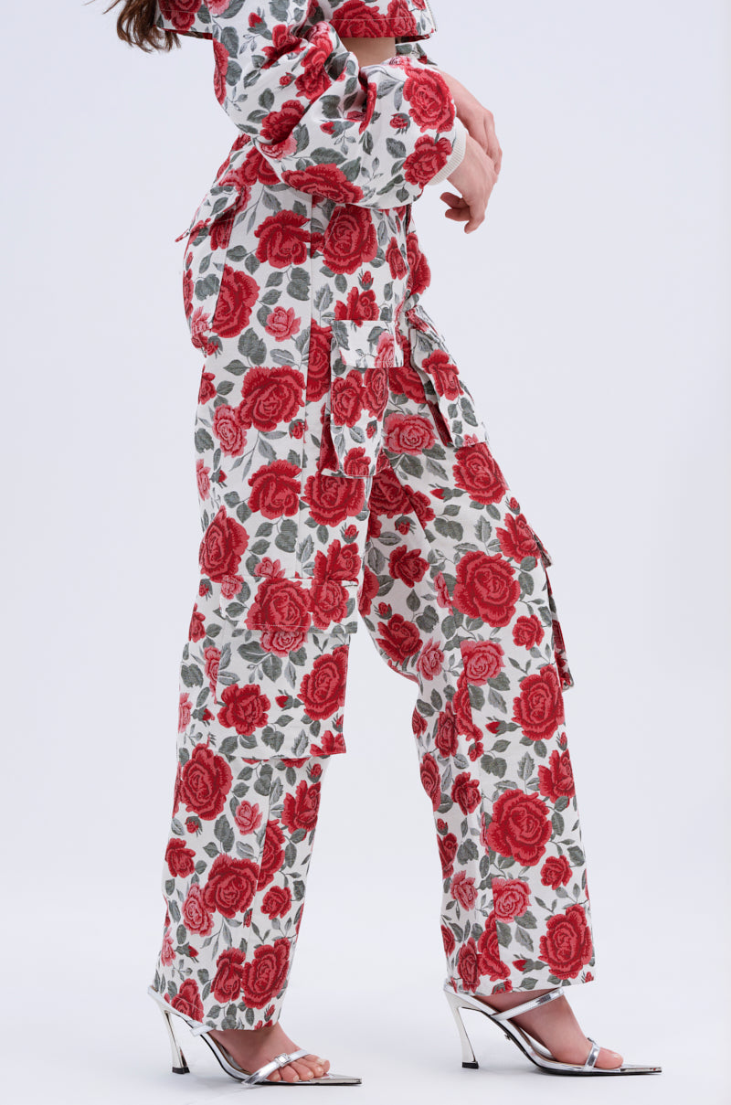 A person stands against a plain light background wearing the FULL OF ROSES TROUSER with a red rose floral pattern, a matching long-sleeve top, and silver high-heeled sandals.