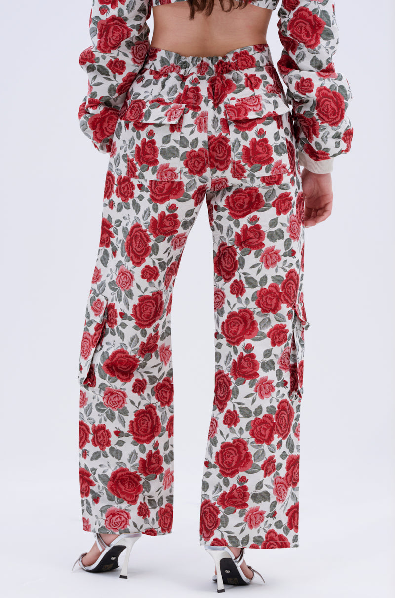 A person stands facing away, wearing the FULL OF ROSES TROUSER with a white rose print and wide legs, paired with a matching vibrant floral jacket and white high-heeled sandals against a plain light background.