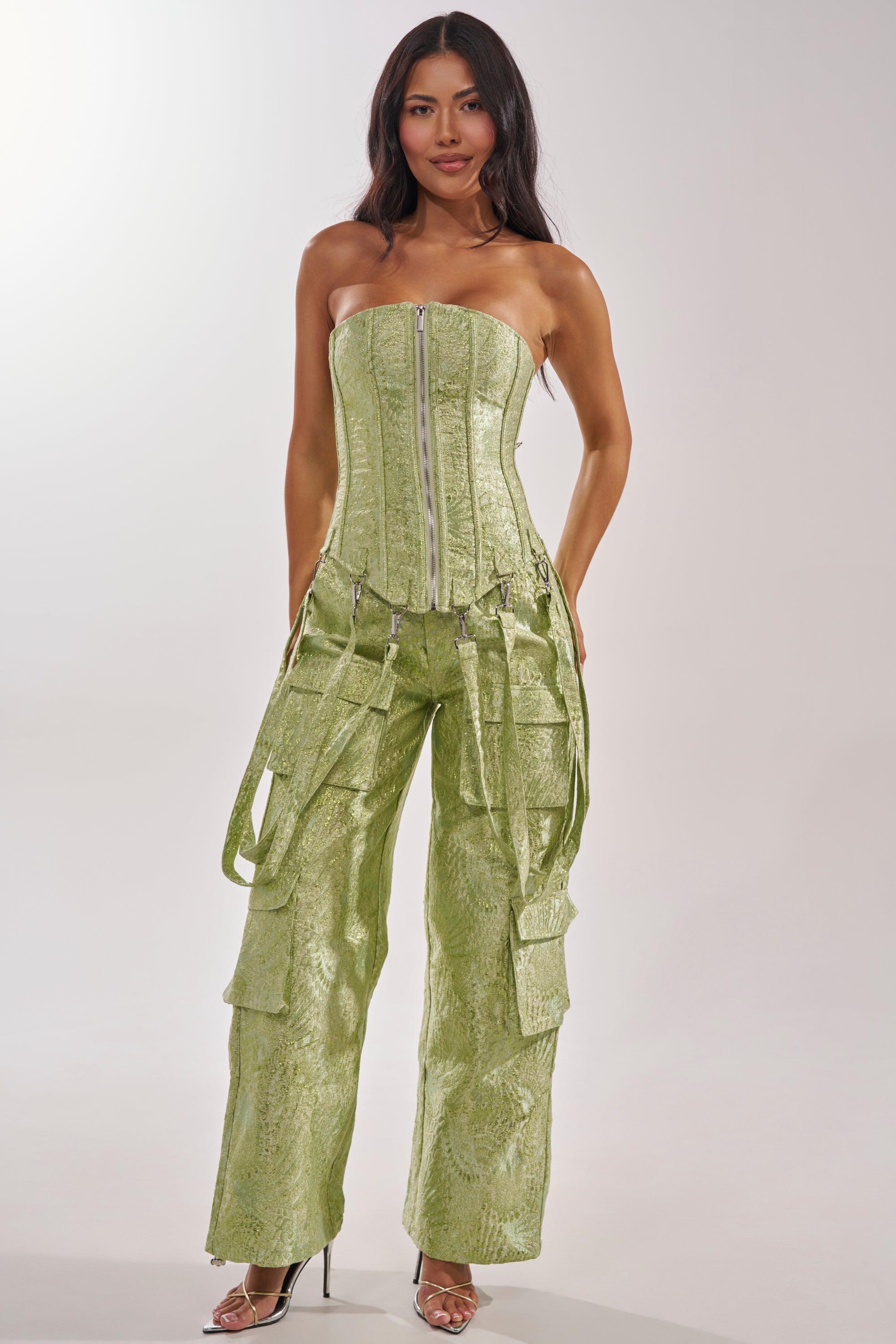 A woman wears the ENVY ME BROCADE CORSET TOP, a strapless green brocade piece, paired with matching high-waisted cargo pants with pockets and straps, styled with clear heels against a plain background. 1