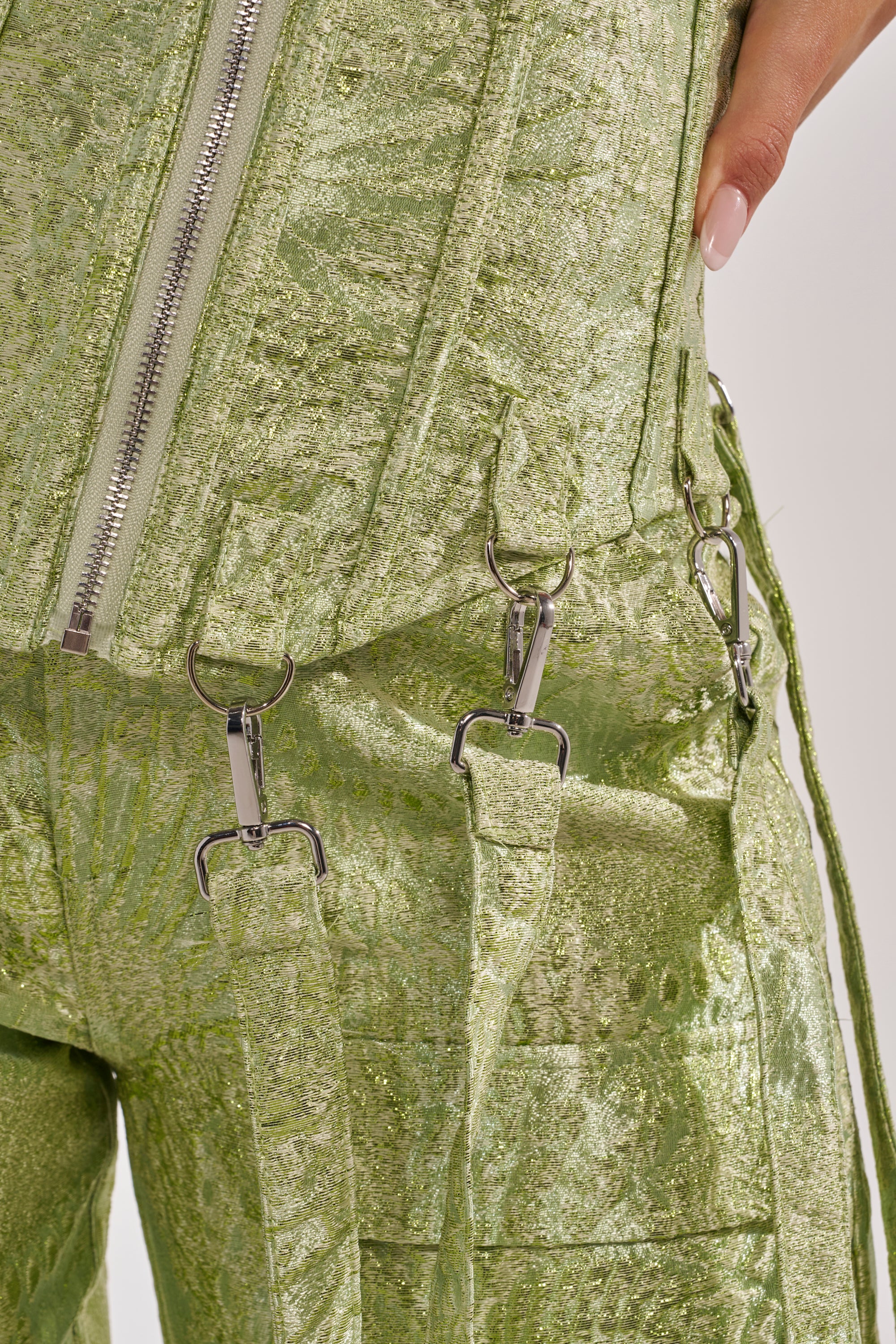 Close-up of a person in shiny, textured light green pants with silver zippers and metal hooks. Their manicured hand rests near the waistband, paired with the ENVY ME BROCADE CORSET TOP for a bold nightlife look. 12