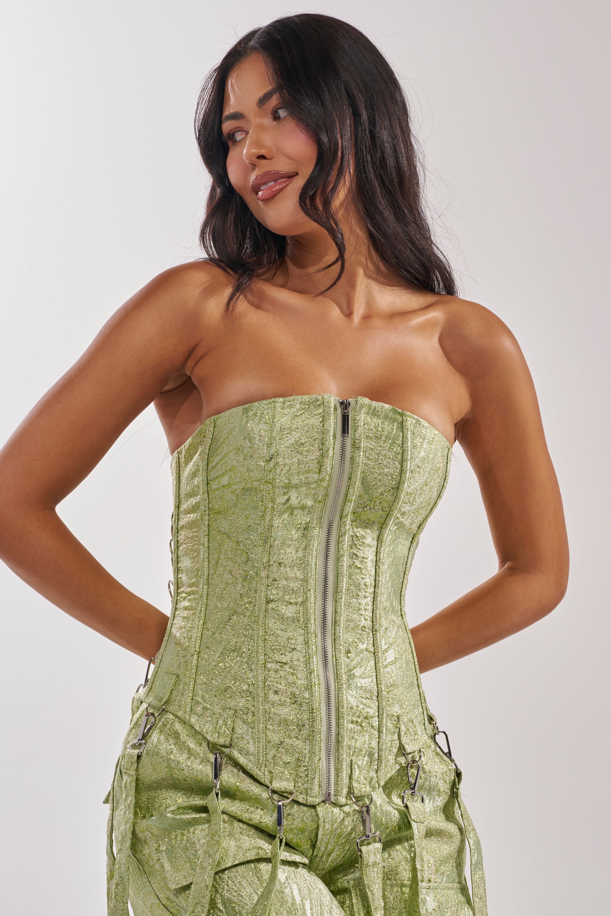A woman with long dark hair wears the ENVY ME BROCADE CORSET TOP, a strapless green brocade piece with a front zipper, paired with matching pants. She stands smiling softly, hands behind her back and looking slightly to the side. 2