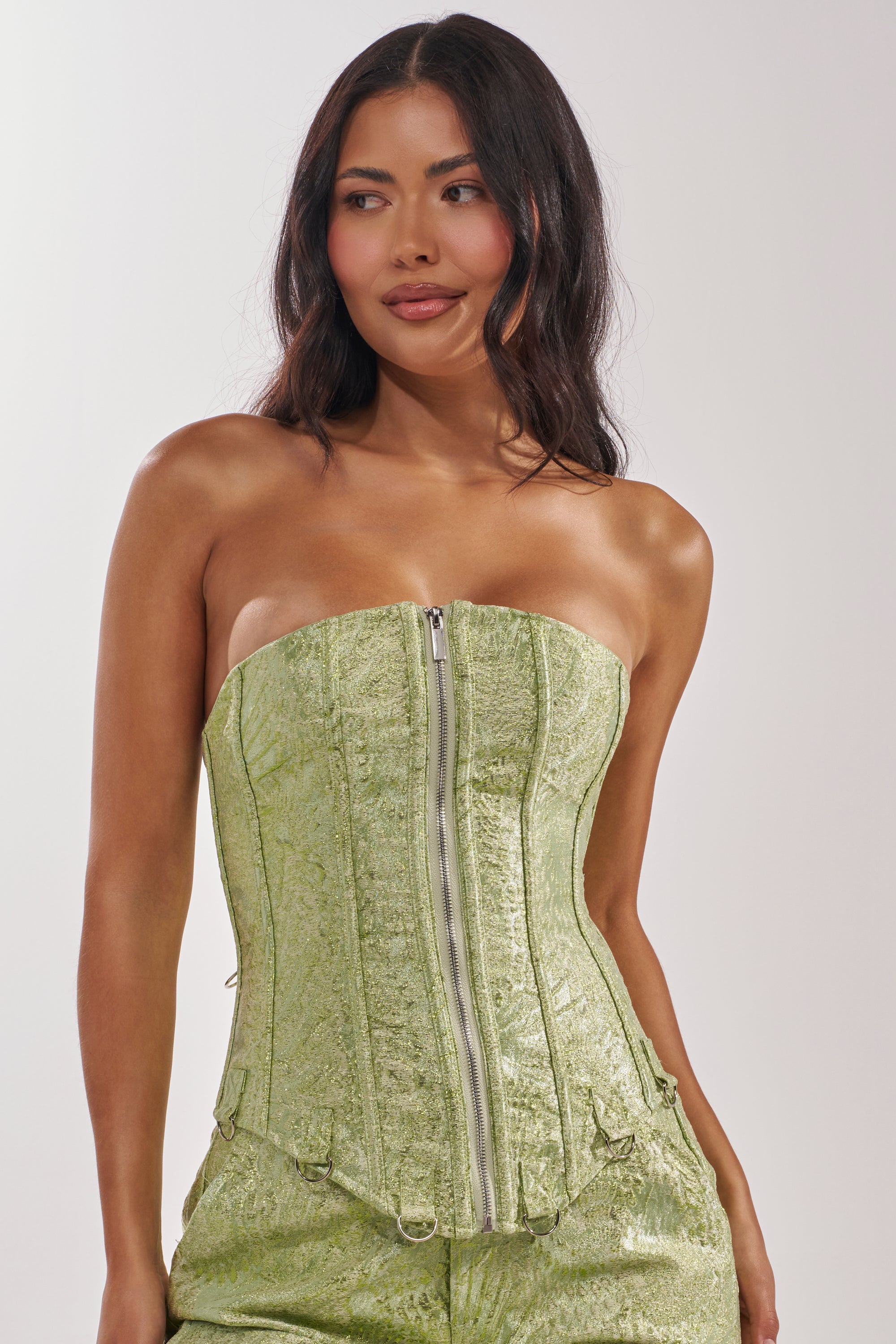 A woman with long, dark hair models the ENVY ME BROCADE CORSET TOP with a front zipper and matching pants, creating an eye-catching nightlife look against a plain background. 7