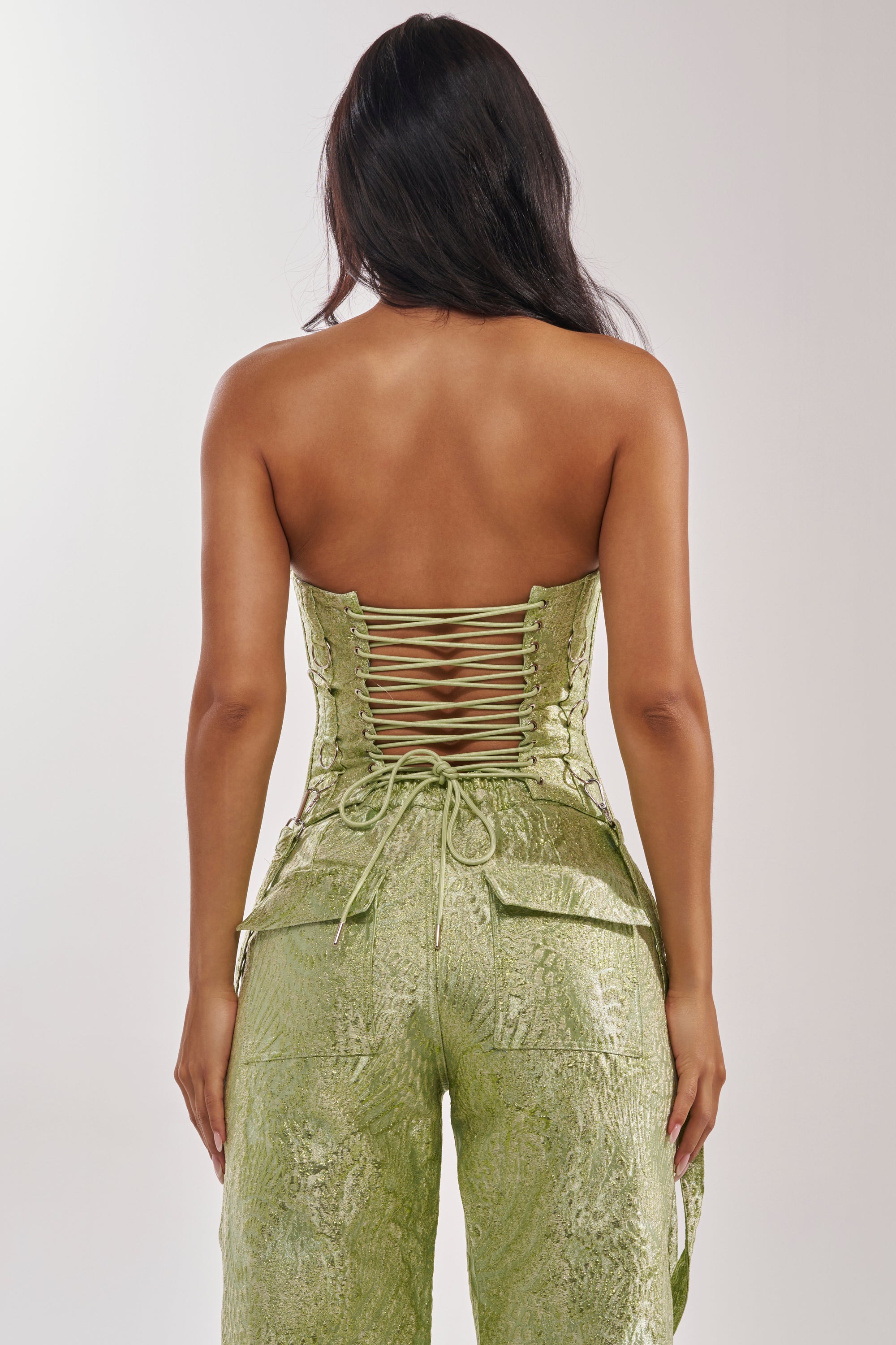Woman shown from the back wearing the ENVY ME BROCADE CORSET TOP in textured green with lace-up detailing and matching high-waisted pants. Her long dark hair cascades down her back, creating a stunning nightlife look against a plain background. 9