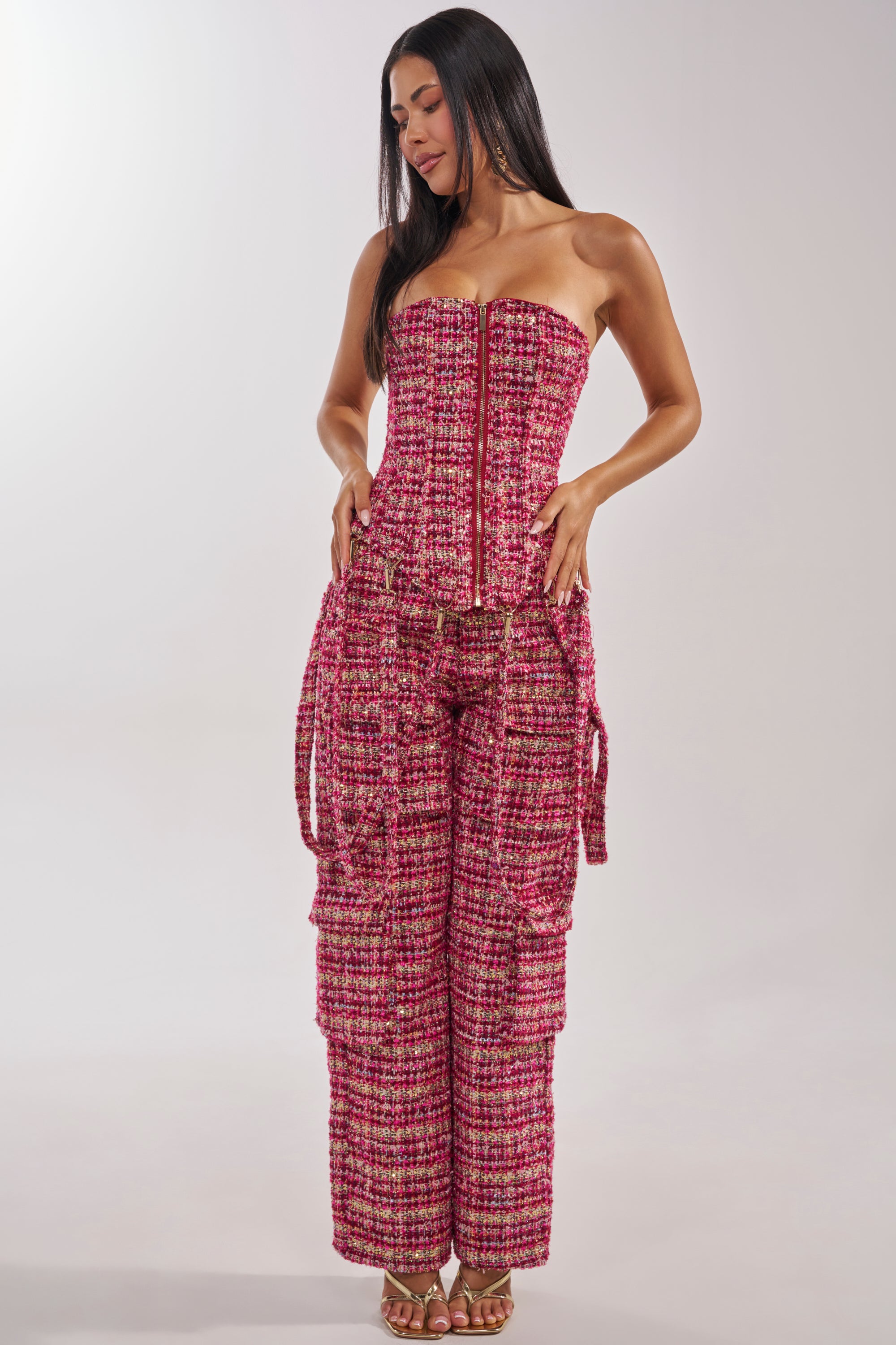 A woman with long dark hair, smiling slightly and looking down, wears the ITS TIME TWEED CORSET—a strapless pink, red, and white tweed corset top with metallic threads, front zipper, and matching wide-leg pants. 9
