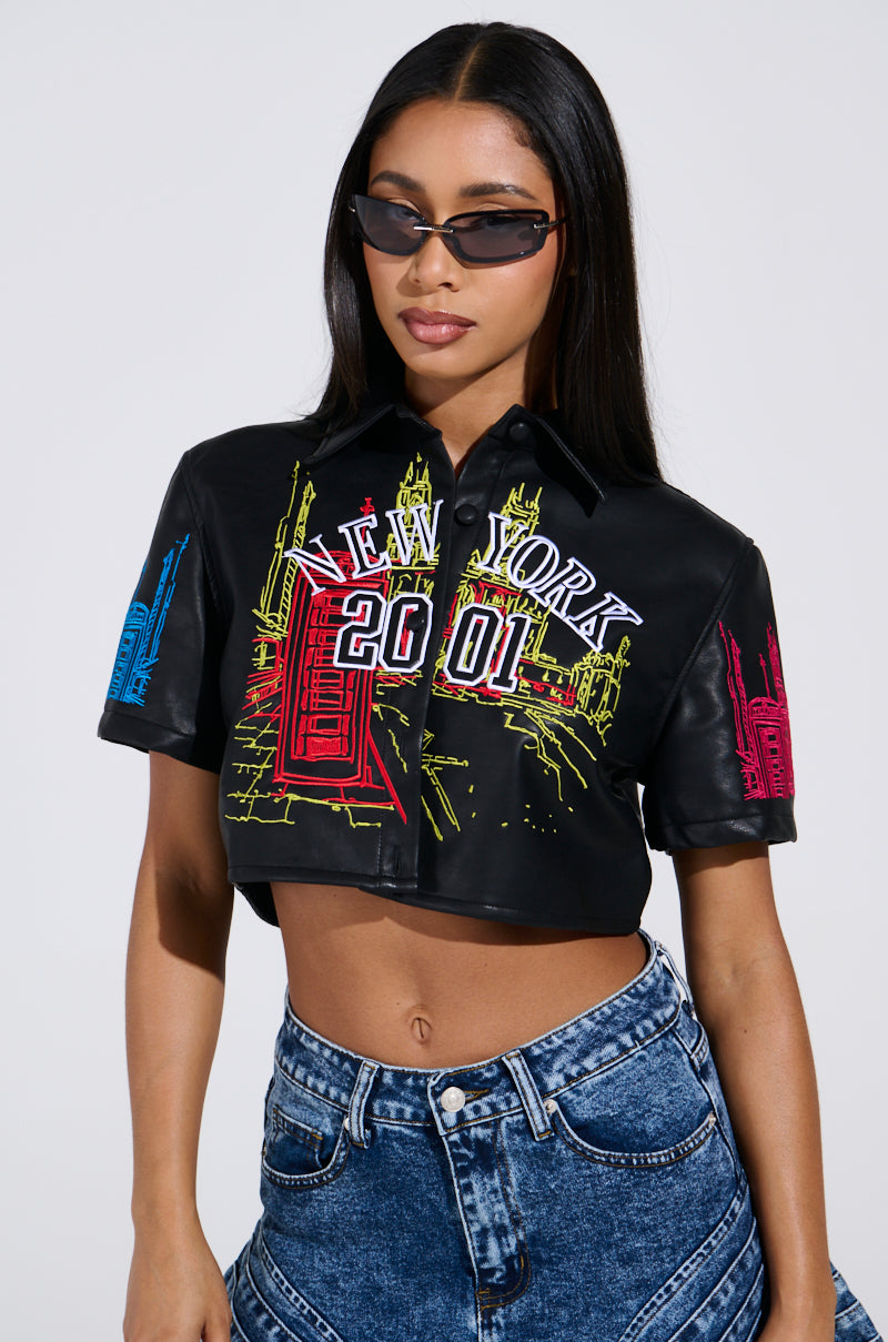 A person wearing sunglasses and denim jeans confidently poses in a black crop top with colorful New York graphics. The top features "New York 2001" with vibrant building outlines against a plain background, highlighting their chic ensemble.