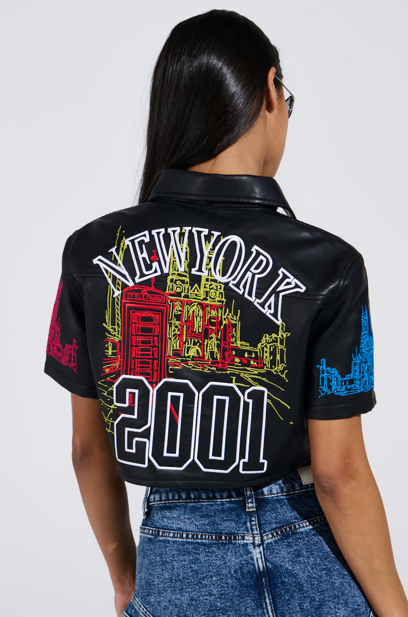 Someone with long hair sports the MAD CITY FAUX LEATHER SHORT SLEEVE BUTTON DOWN SHIRT, showcasing bold white "NEW YORK" and "2001" text with vibrant red, yellow, and blue cityscape designs. The cropped silhouette stylishly complements their blue jeans.