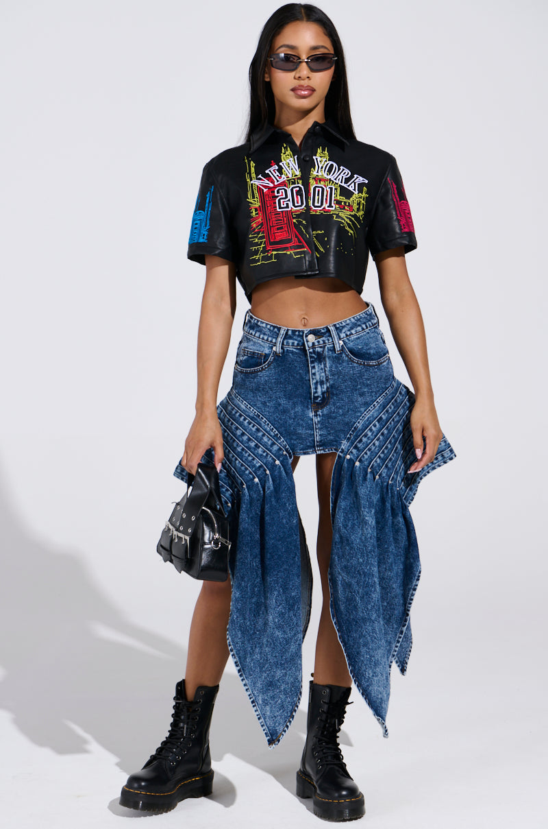 A woman poses confidently wearing a black crop top with colorful graphics, paired with a distinctive blue denim skirt. Complementing her look are black boots and a small faux leather handbag. Stylish sunglasses complete her ensemble as she stands against a plain background.
