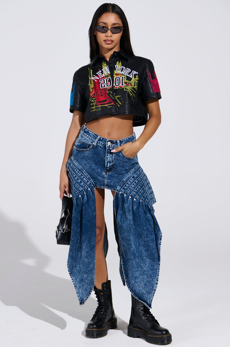 Wearing a chic vibe, a person stands confidently in sleek sunglasses and the MAD CITY faux leather short sleeve button-down shirt. They sport unique wide-legged pleated denim pants with slits, embroidered text, stylish black boots, and a sleek black handbag against a plain backdrop.