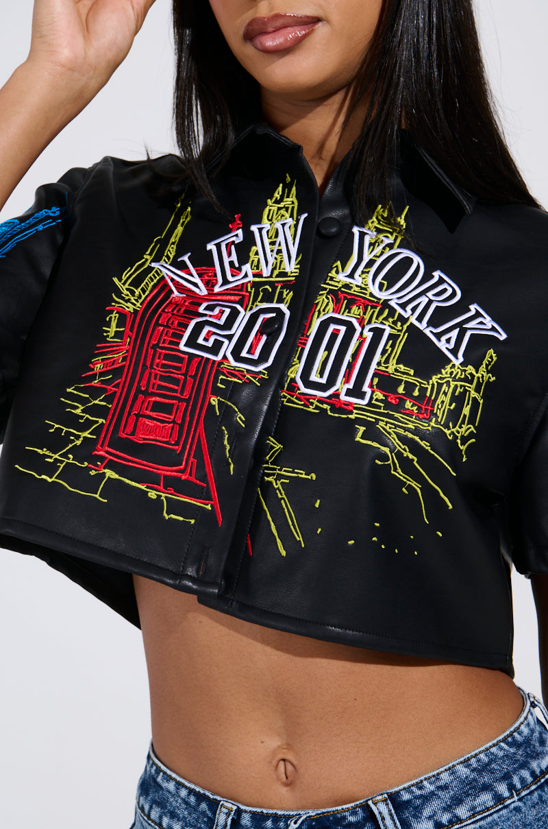 A person is wearing the MAD CITY FAUX LEATHER SHORT SLEEVE BUTTON DOWN SHIRT, featuring "New York 2001" embroidery and a red architectural design on the front. Jeans are visible beneath, with one hand lifted near their face.